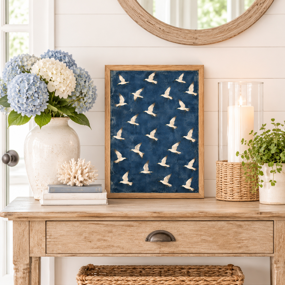 Coastal bird artwork in a natural wood frame displayed on a wooden entryway table with blue hydrangeas, white coral, and neutral beach house accessories.