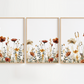 Three framed vertical floral art prints with a watercolor-style wildflower design in muted orange, cream, yellow, and olive tones, displayed side by side on a white wall.