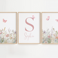 Three framed botanical wall art prints hang on a white wall. The center print features a large pink letter S and the name Sophia, while the left and right prints show pink butterflies above pastel wildflowers in light wood frames.