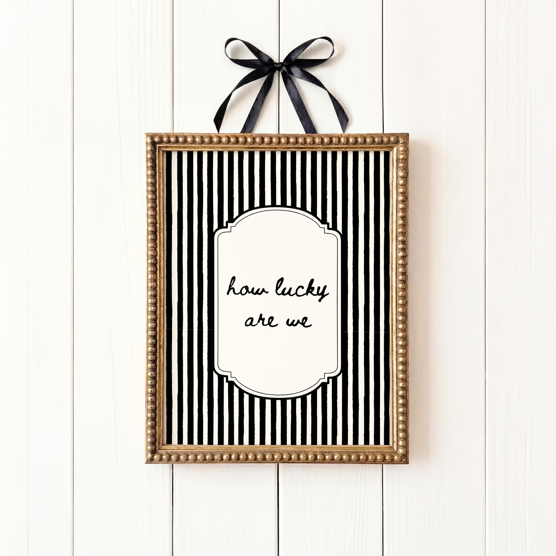 Framed black and white striped print with 'how lucky are we' text on a white wooden background.