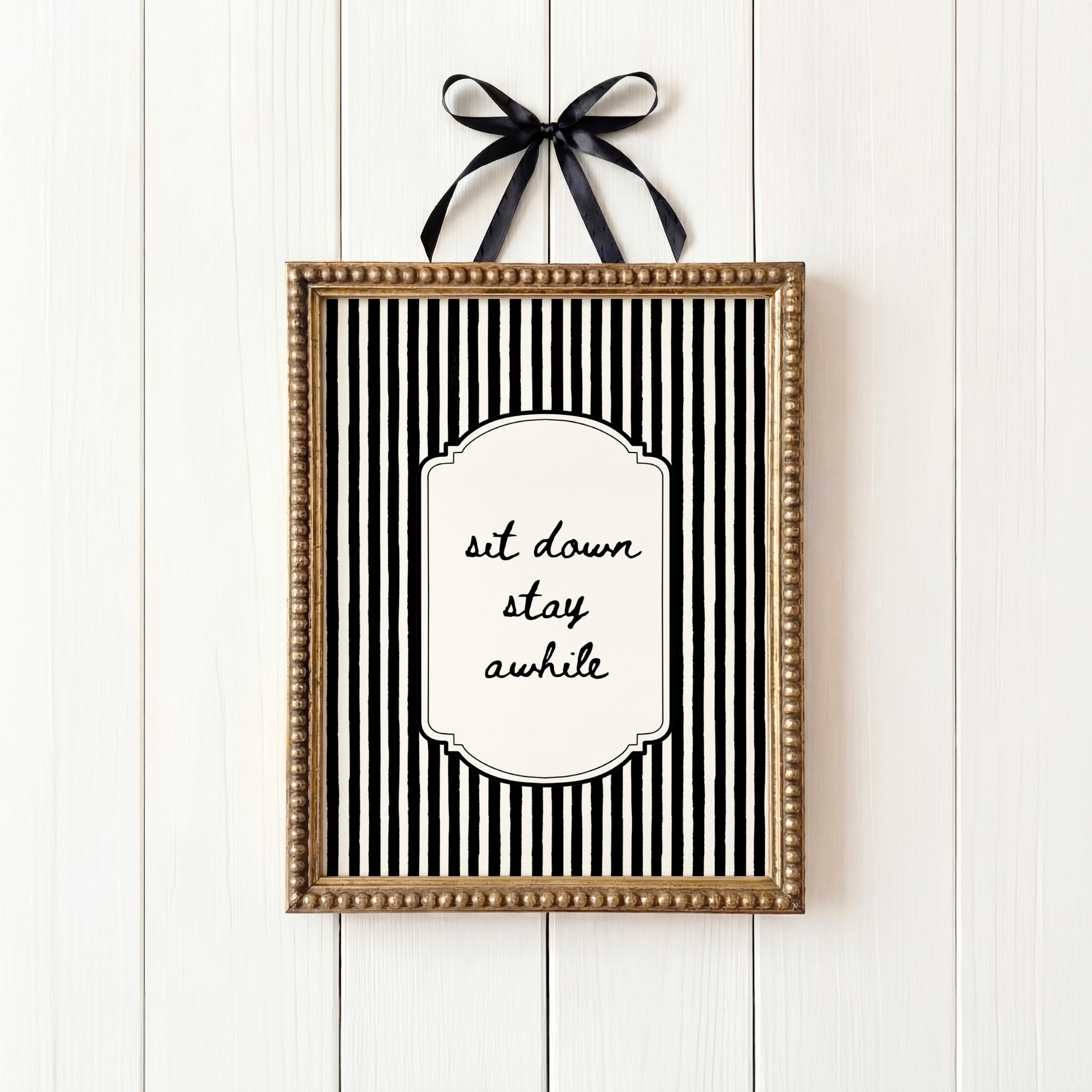 Framed black and white striped print with 'sit down stay awhile' text on a white wooden background.