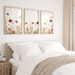 Three vertical framed floral prints with wildflower illustrations hang above a neatly made bed in a bright, neutral bedroom. The frames have light wood borders, and the bedding is white with a beige knit throw blanket.